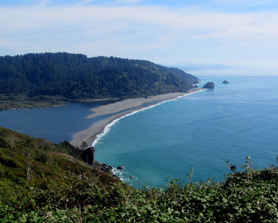 A scenic overlook view of the Klamath River flowing into the Pacific Ocean in the North Coast region of California, completing its watery path through the Del Norte Coast Redwoods State Park and Redwood National Park. Activities include fishing, bird watching and walking along the beach. (Image created by USA Patriotism! from U.S. National Park Service photo.)