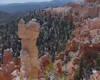 A captivating scenic view of a mysterious, strange shaped hoodoo formation along a trail near the Hat Shop with a small group of other narrow, eroded, orange-colored pinnacles topped by wider, grey boulders, far below the edge of the plateau in the north section of Bryce Canyon National Park. Beyond to the horizon is mostly wooded land bordered by a mountain range. The trail to Hat Shop is one of Bryce’s least-traveled day hikes, complete with great views and a point of interest as its destination. (Image created by USA Patriotism! from U.S. National Park Service photo.)